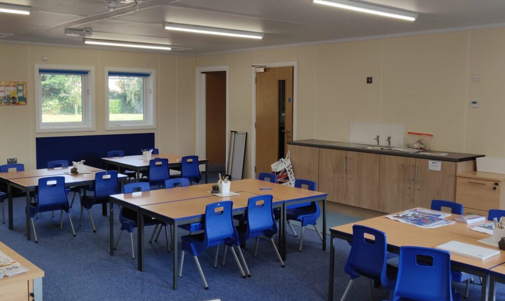 Classroom with blue chairs and rectangular wooden tables arranged in groups. Sinks and storage cabinets are against the wall with a door. Two windows and a whiteboard are visible.