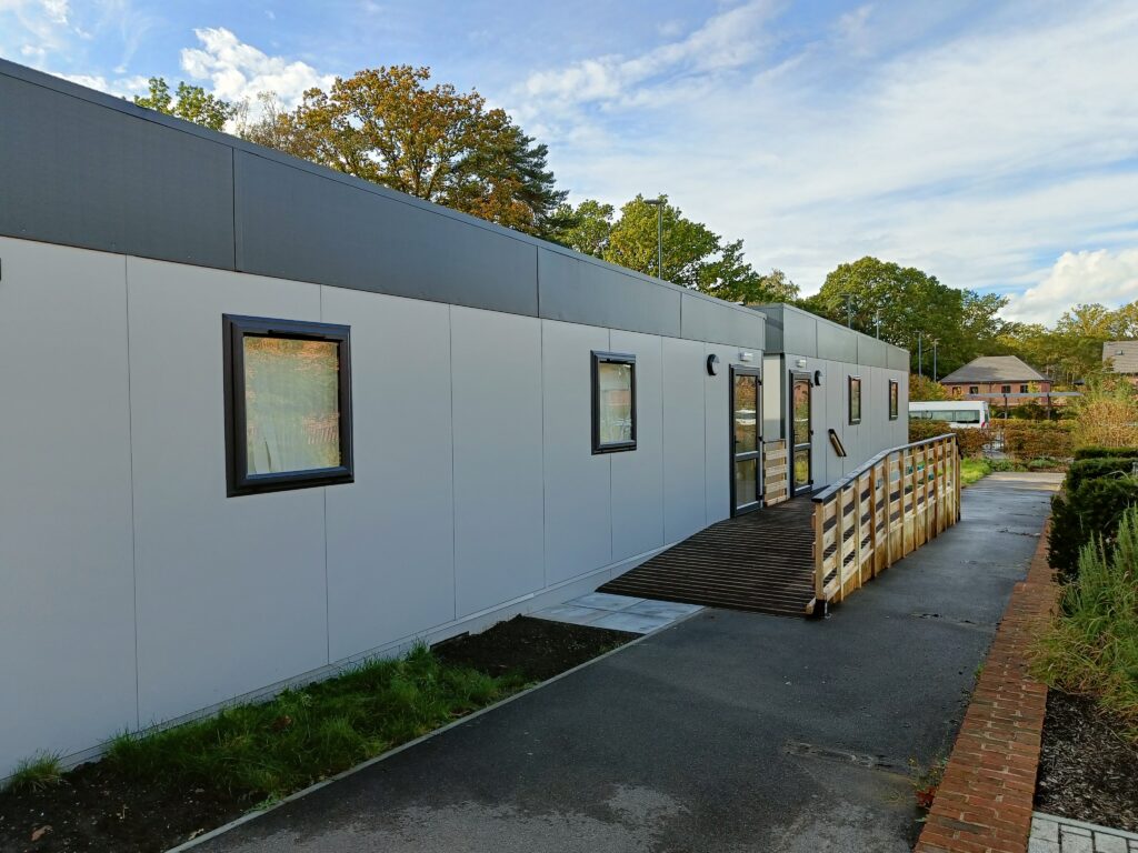 A long, rectangular white building with several windows and a wooden wheelchair ramp, located in an outdoor area with greenery and neighboring buildings in the background. Trees and blue sky visible.