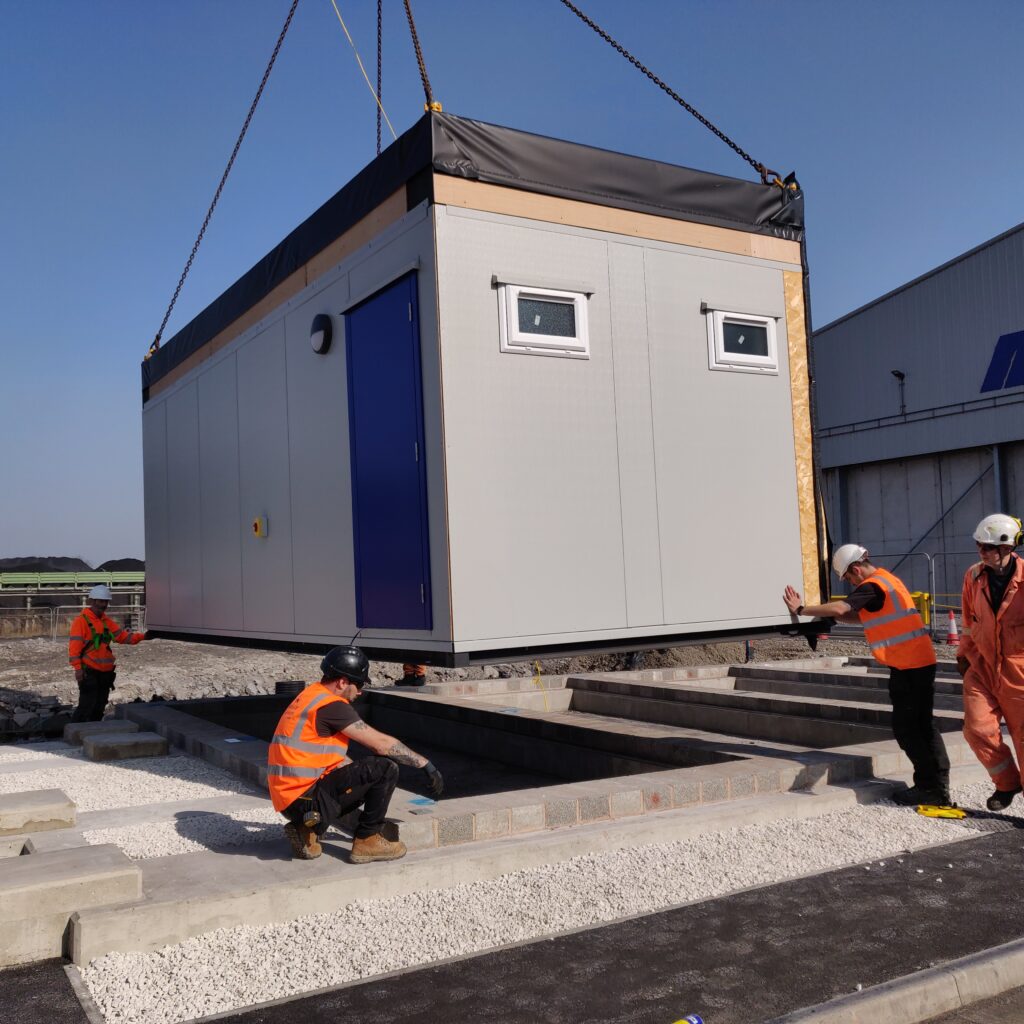 A modular building is being lifted by a crane onto a construction site, with four workers in orange safety gear guiding its placement. The area has gravel, cement blocks, and a nearby building.