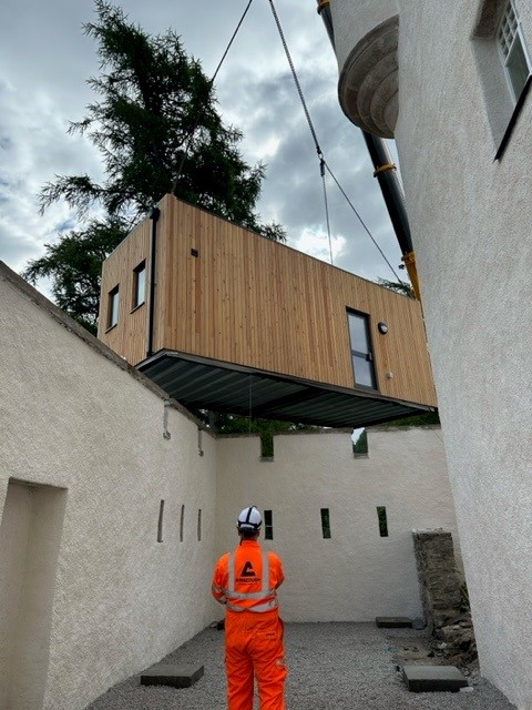 A modular wooden building section is being lifted by a crane over white stucco walls, with a construction worker in a helmet observing the process below.