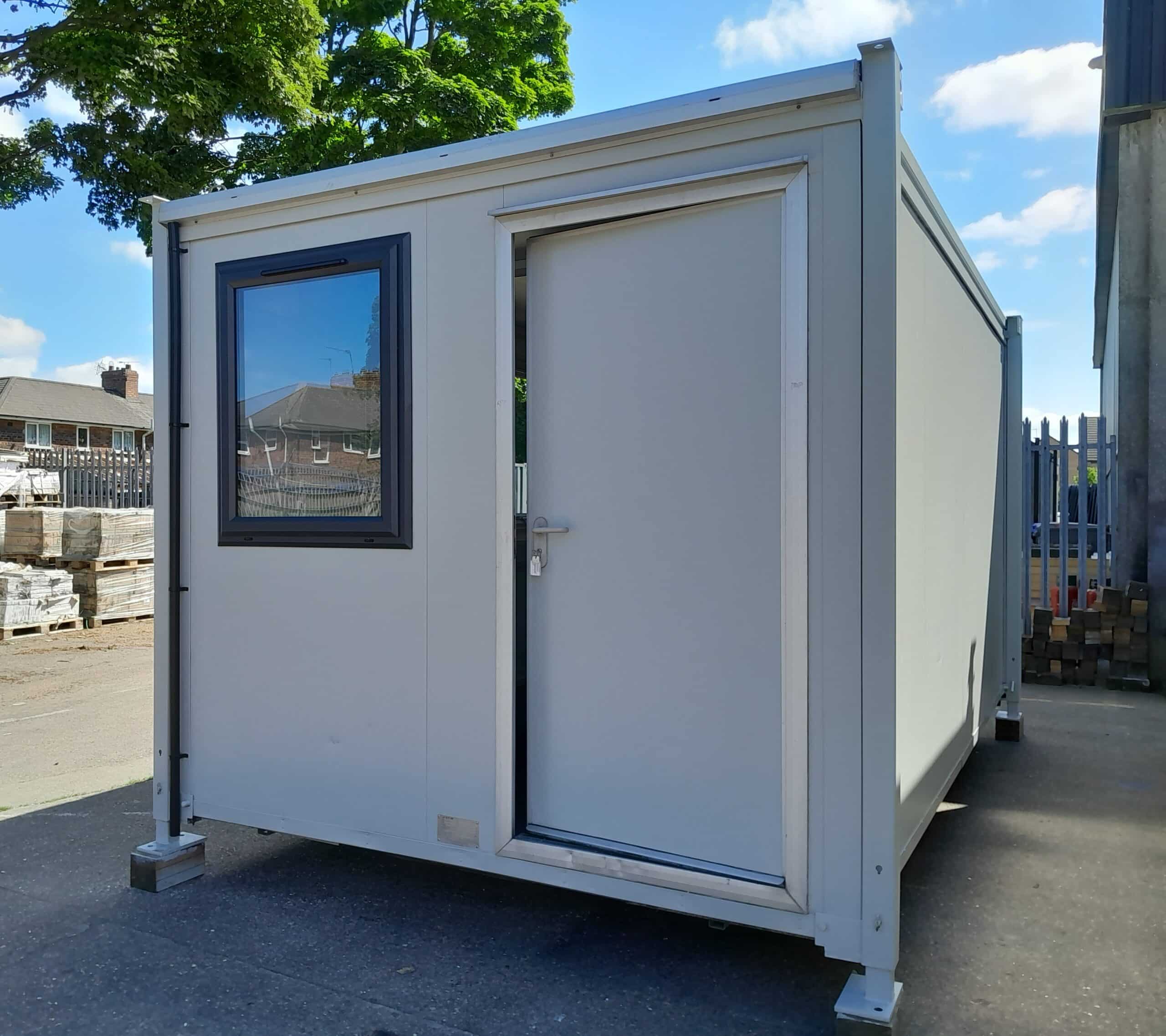 A small, grey portable cabin with a window and partially open door, positioned on a paved outdoor area with stacked pallets and a fence in the background.